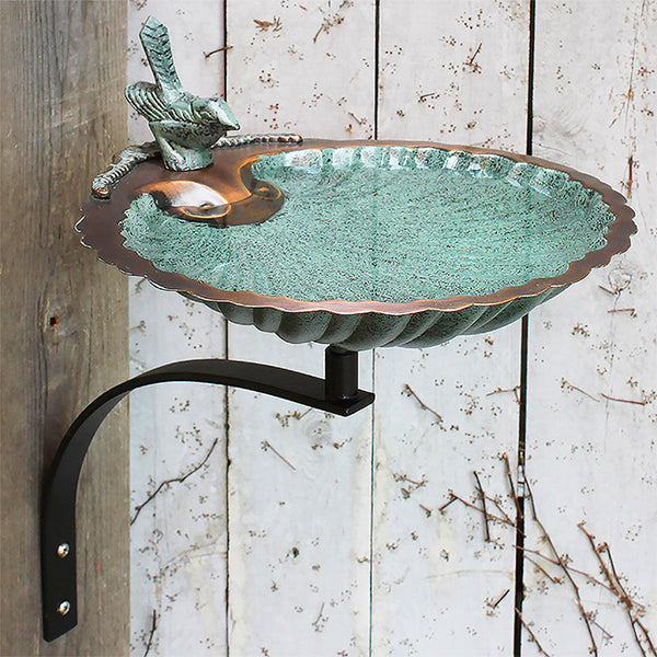 Our Verdigris Scalloped Shell Birdbath with Black Metal Wall Bracket  is crafted from aluminum in the shape of a seashell. It is a beautiful accent piece for any style outdoor space. The black metal wall bracket creates a beautiful contract to the bowl. A metal bird figurine rests on the edge of this basin, and its lifted tail and forward gaze bring height and personality to the piece