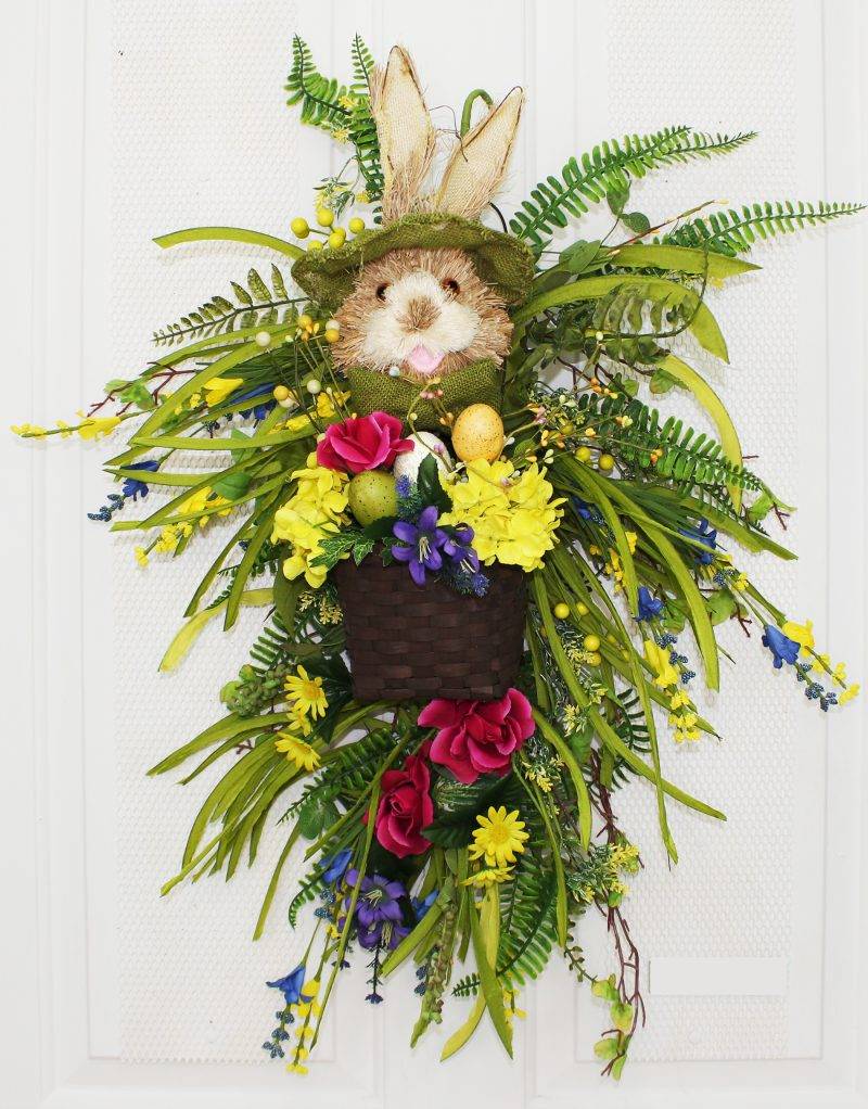 Our Bowtie Bunny With Basket Easter Spring Teardrop Front Door Wreath is 23” in length and features colorful silk flowers as well as a festive bunny rabbit all dressed up with green brim hat and matching bowtie and holding a basket full of flowers and Easter eggs and surrounded with lots of wispy greens and ferns