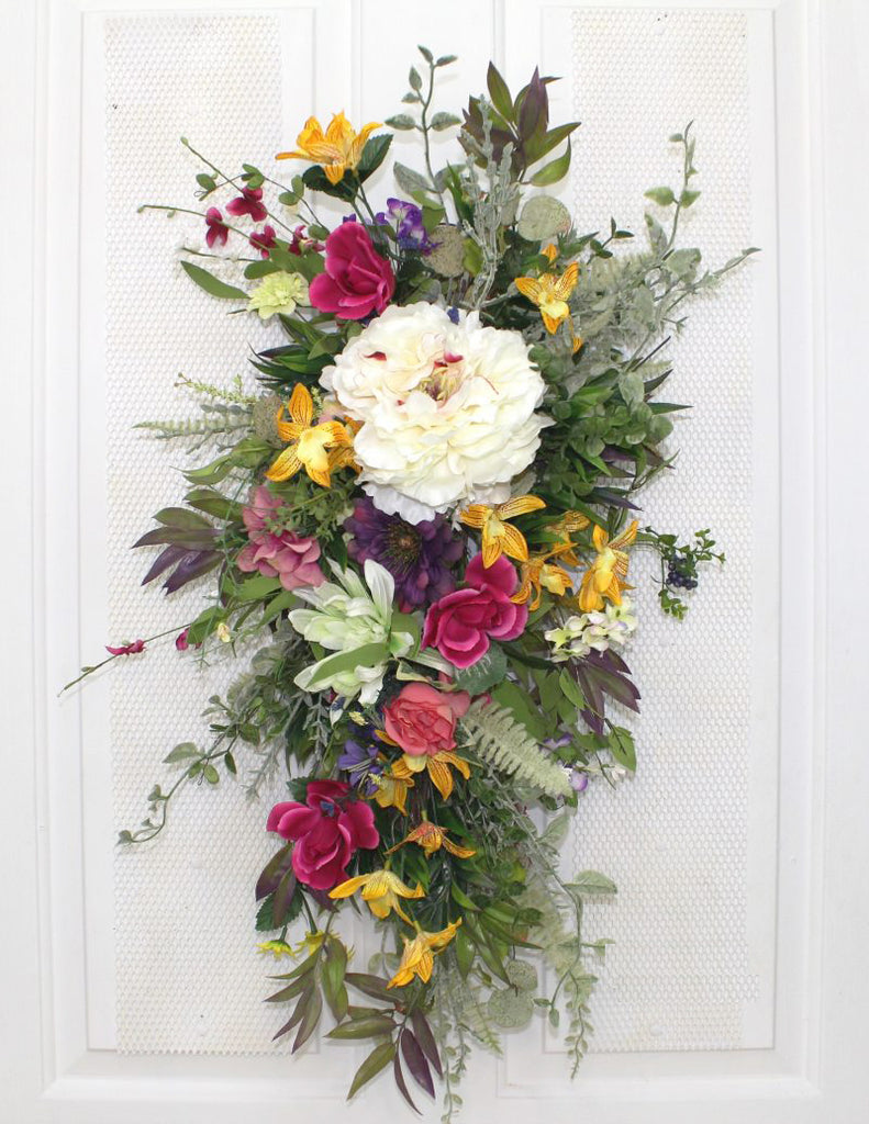This beautifully handcrafted front door wreath is so elegant and stylish that you can display it all year long. This custom made to order decorative teardrop wreath measures 24” in length and features a large peony bloom, surrounded with a full array of colorful flowers in shades of pink, along with scatterings of yellow and purple flowers and lots of greenery .